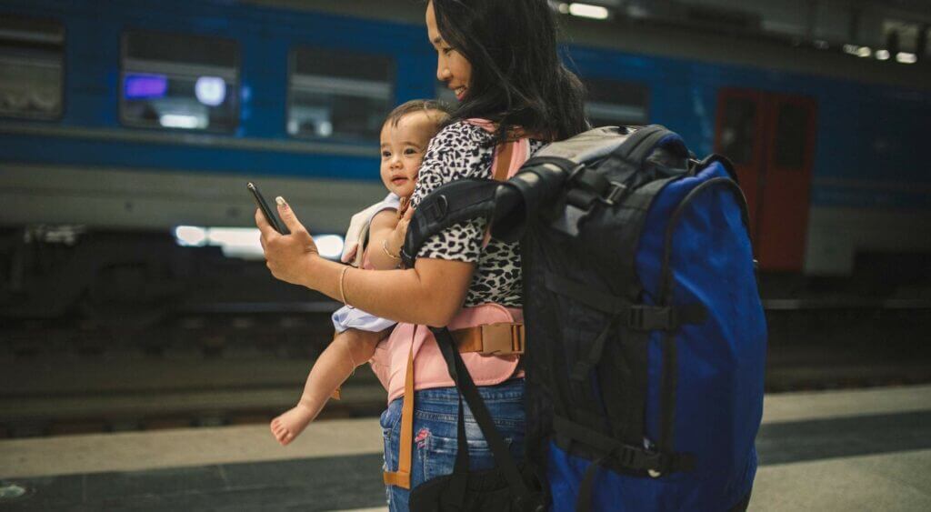 Travel nurse with baby at train station, wearing backpack, using phone; exploring travel nursing with children.
