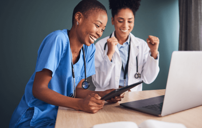 Two healthcare professionals smiling, using laptop and tablet in hospital office, teamwork, medical technology, diverse doctors collaboration.